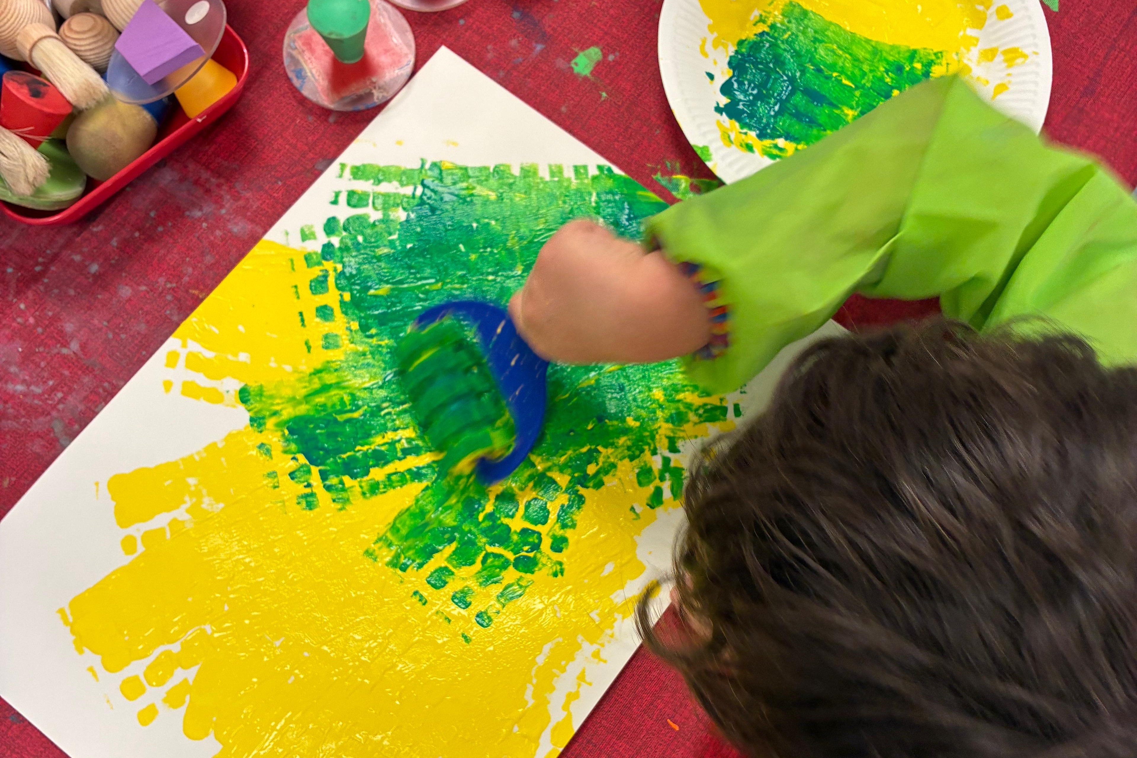 Blick von oben auf ein Kind, das mit einer Strukturrolle bunte Muster auf ein Papier druckt.